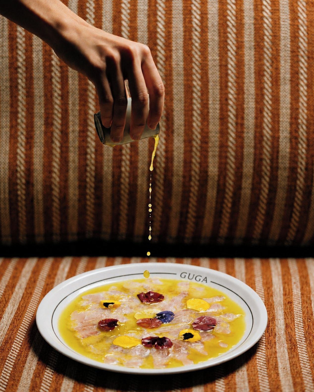 Una mano vertiendo un líquido amarillo sobre un plato con comida y pétalos de flores, etiquetado 'GUGA'.