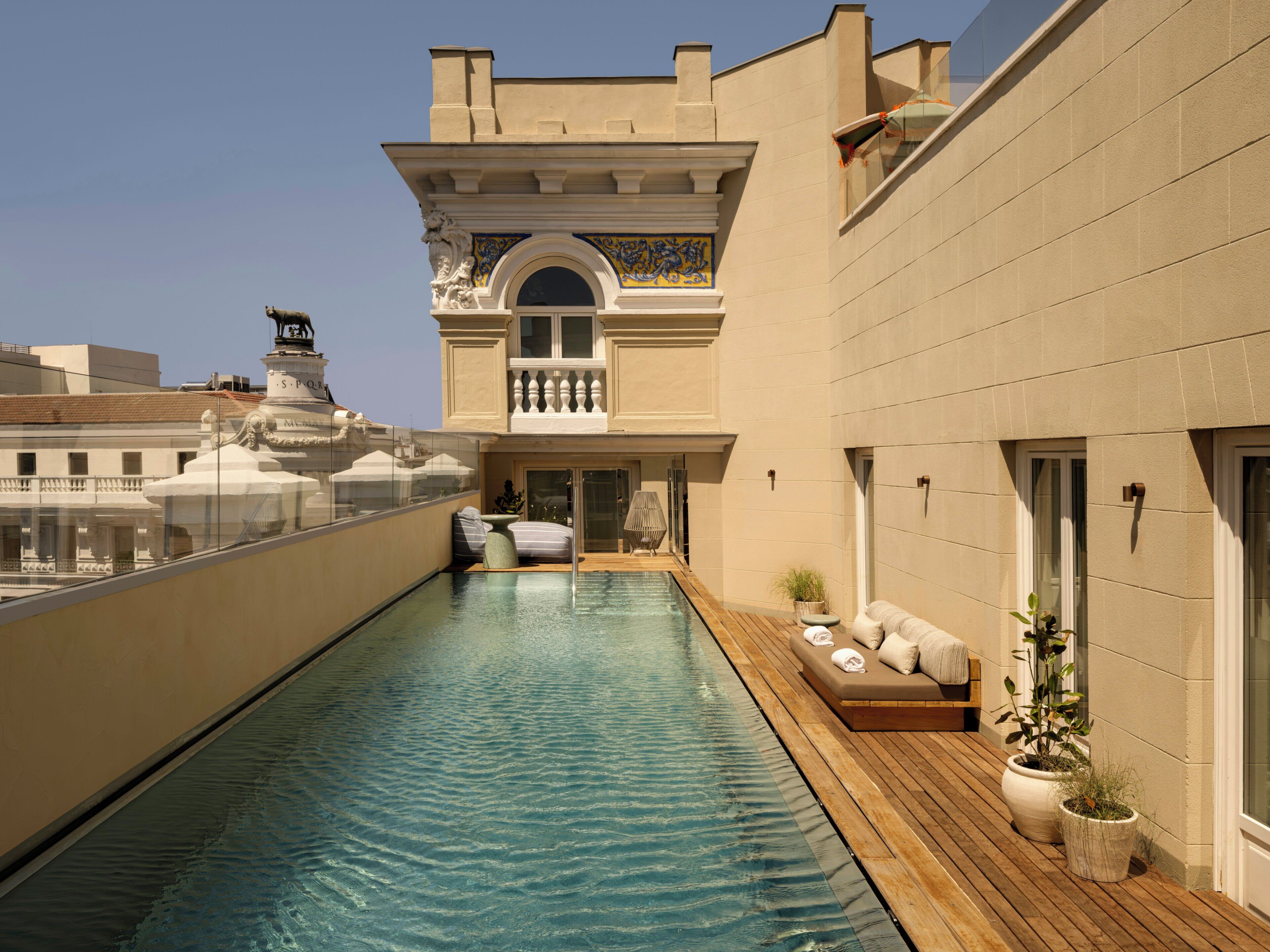 A rooftop pool with a wooden deck, a lounge area with a sofa, and a view of a building with classical architecture.