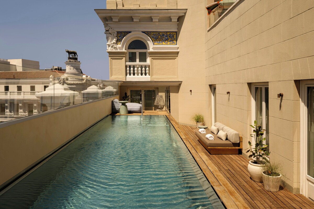 A rooftop pool with a wooden deck, a lounge area with a sofa, and a view of a building with classical architecture.