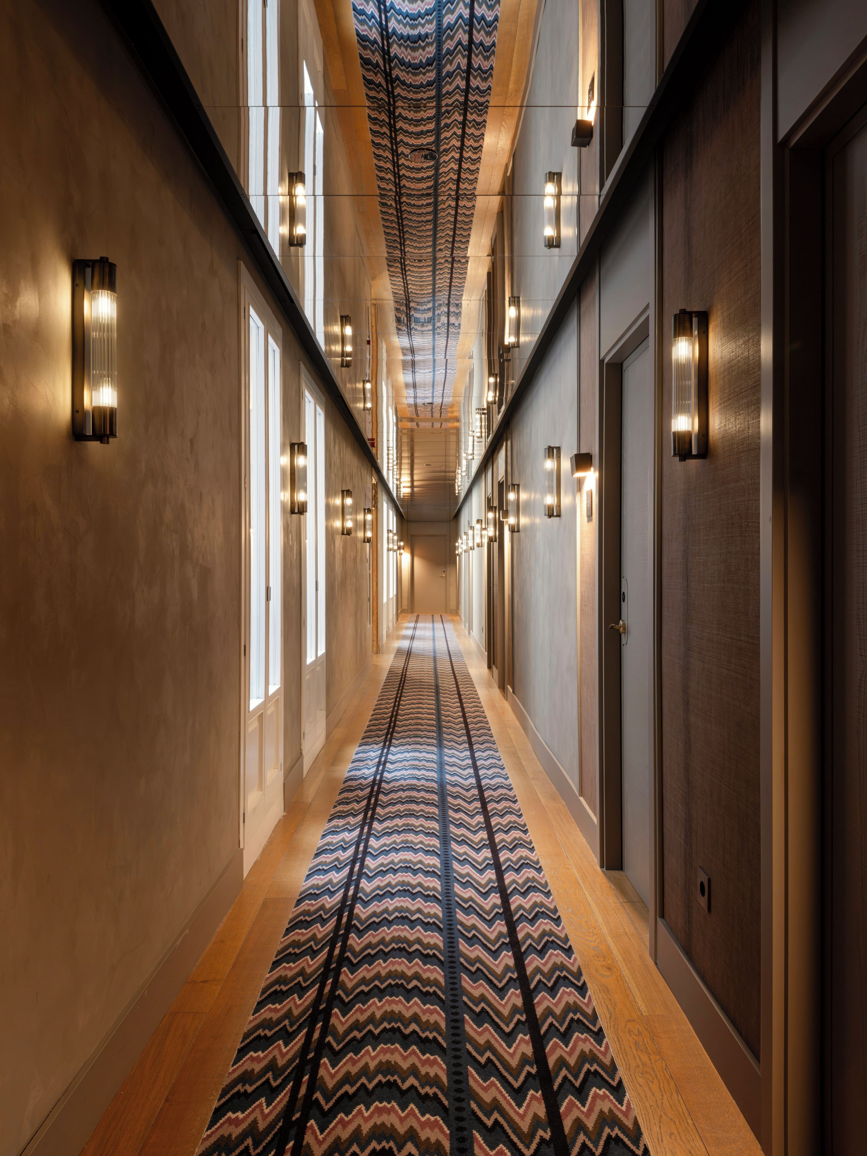 Un pasillo de hotel con alfombra estampada, luces empotradas en la pared y puertas a ambos lados.