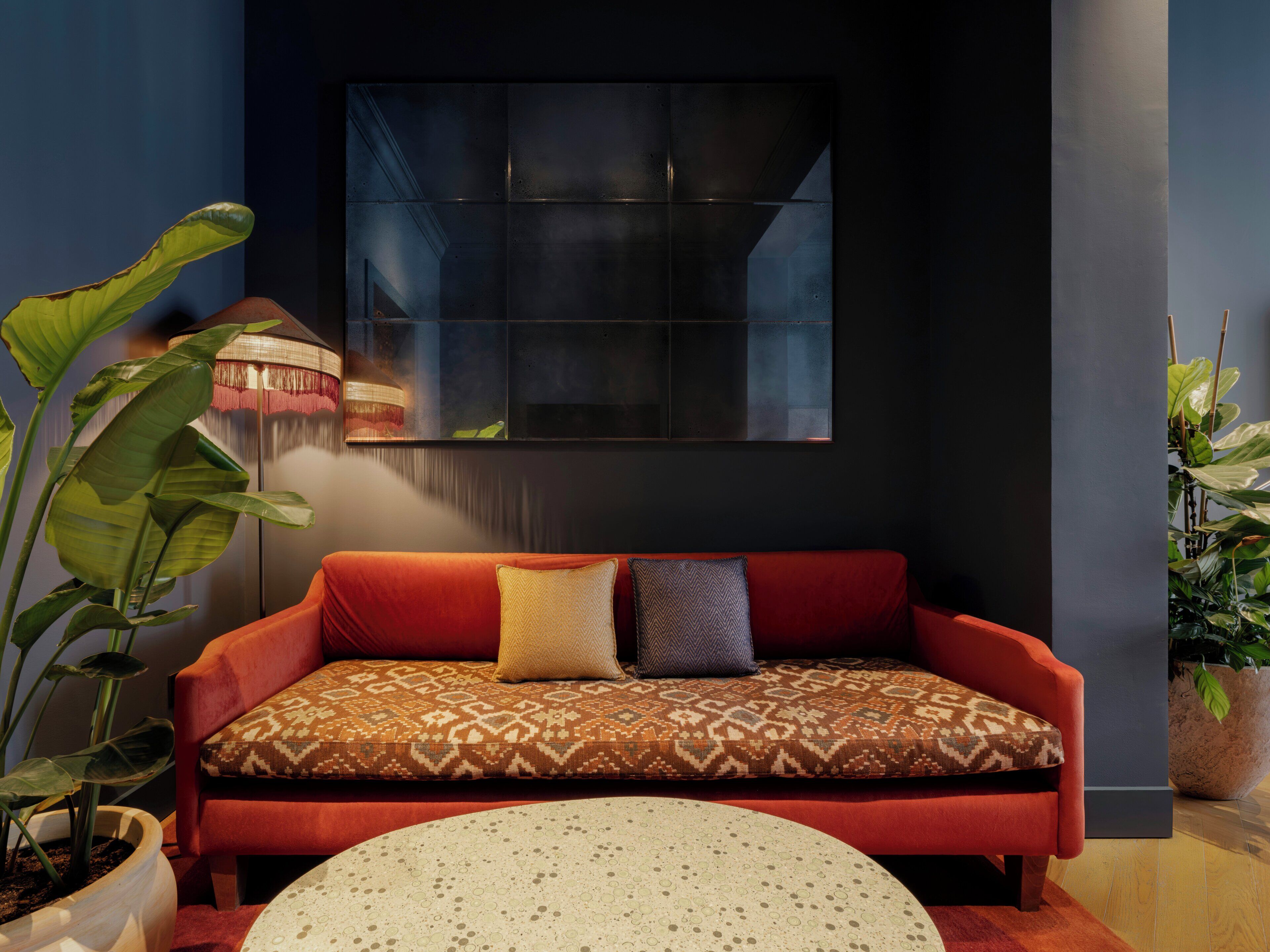Un salon moderne avec un canapé rouge, des coussins à motifs, une table basse ronde et de grandes plantes vertes.
