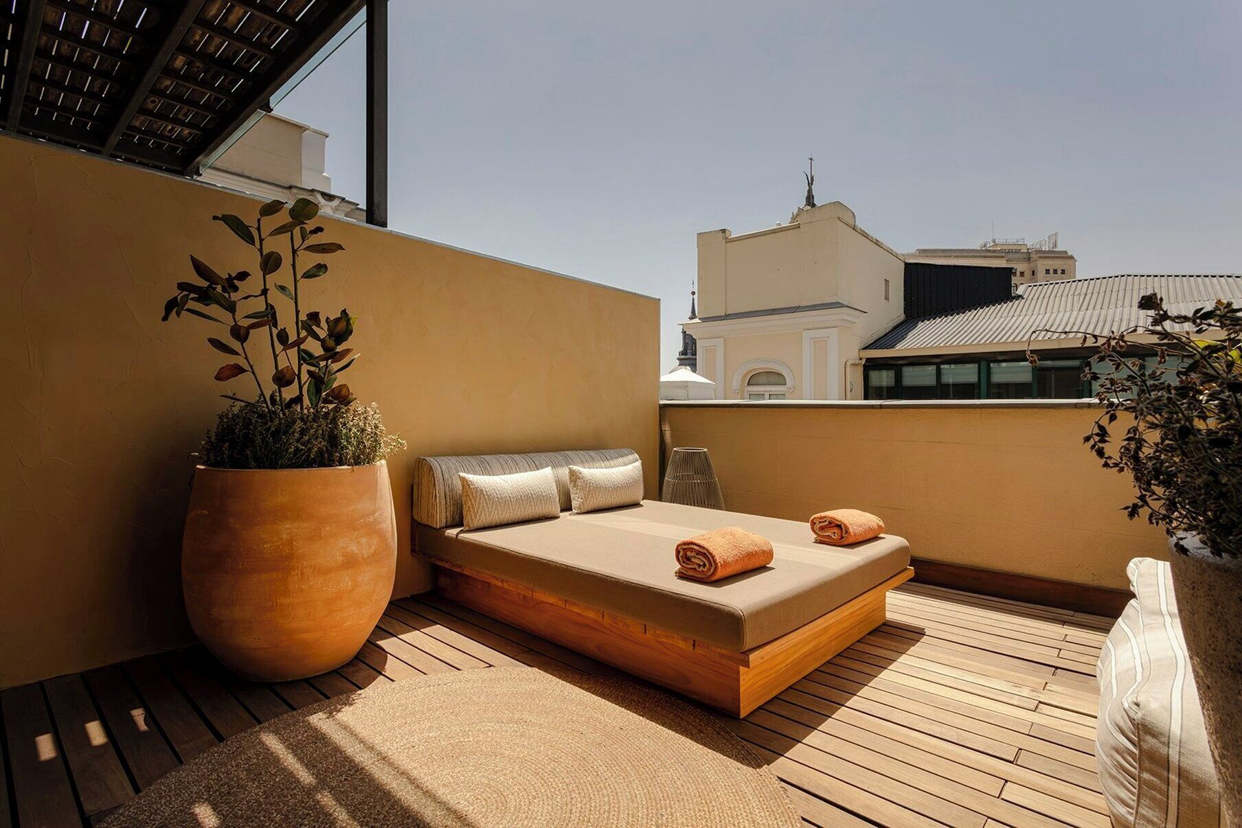 Un piccolo balcone privato con pavimento in legno, un letto singolo con cuscini, una grande pianta in vaso e vista sugli edifici sullo sfondo.