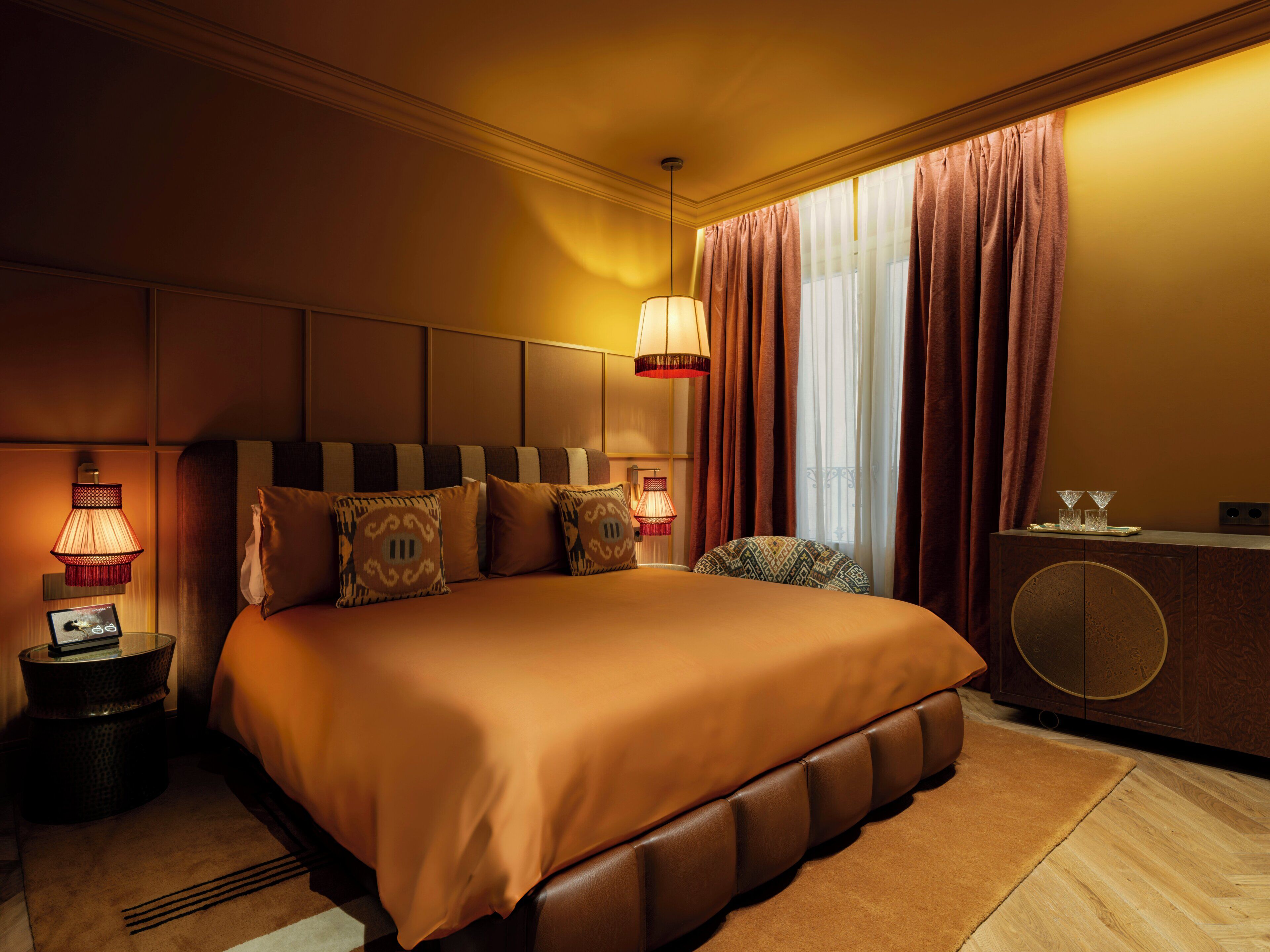 A hotel room with a large bed, a bedside table with a lamp, a small table with a tray of glasses, and a cabinet with a round door.
