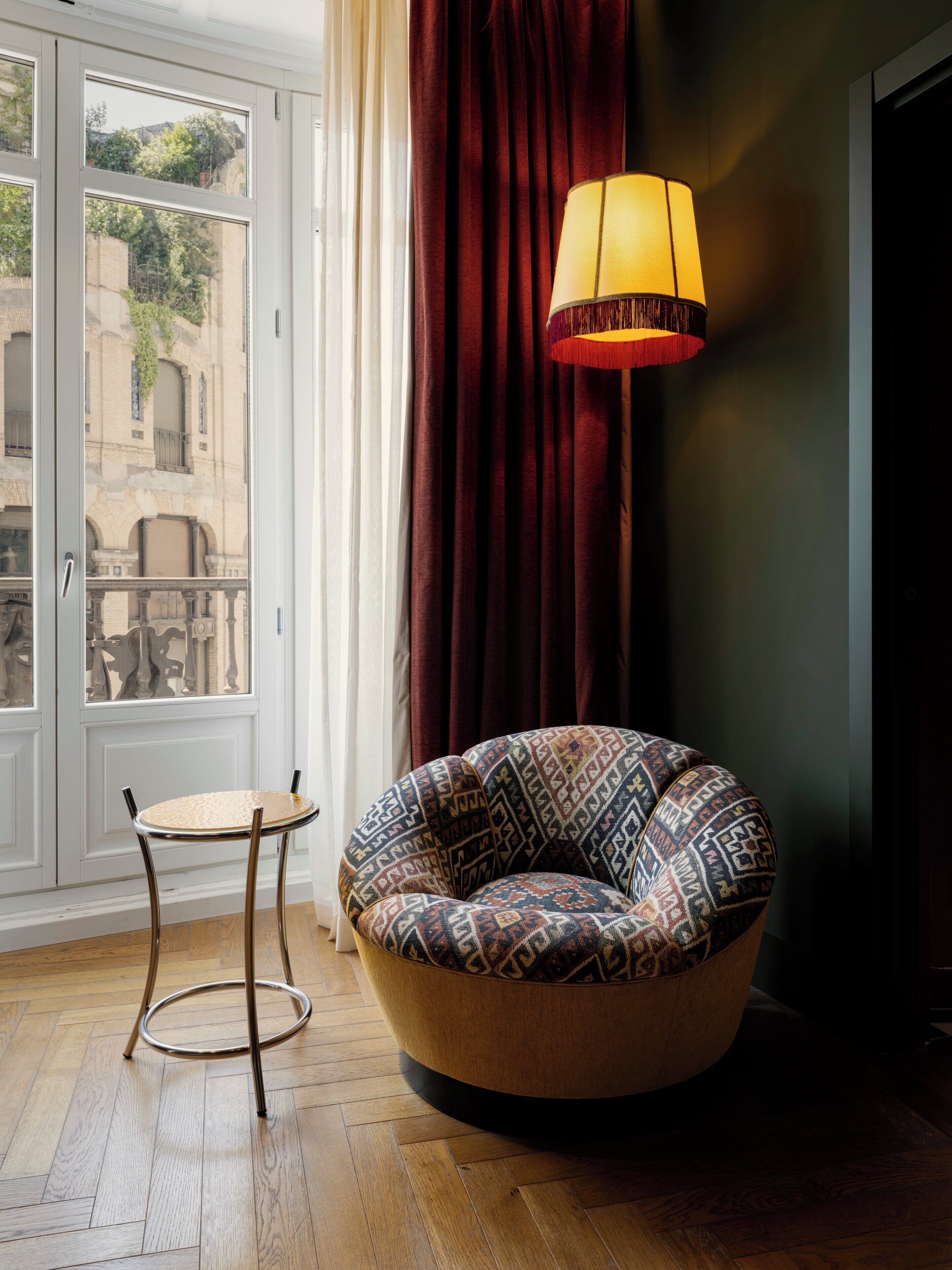 Ein Zimmer mit einem gemusterten Sessel, einem kleinen runden Tisch, einer Stehlampe und Blick auf Gebäude durch ein mit Vorhängen drapiertes Fenster.