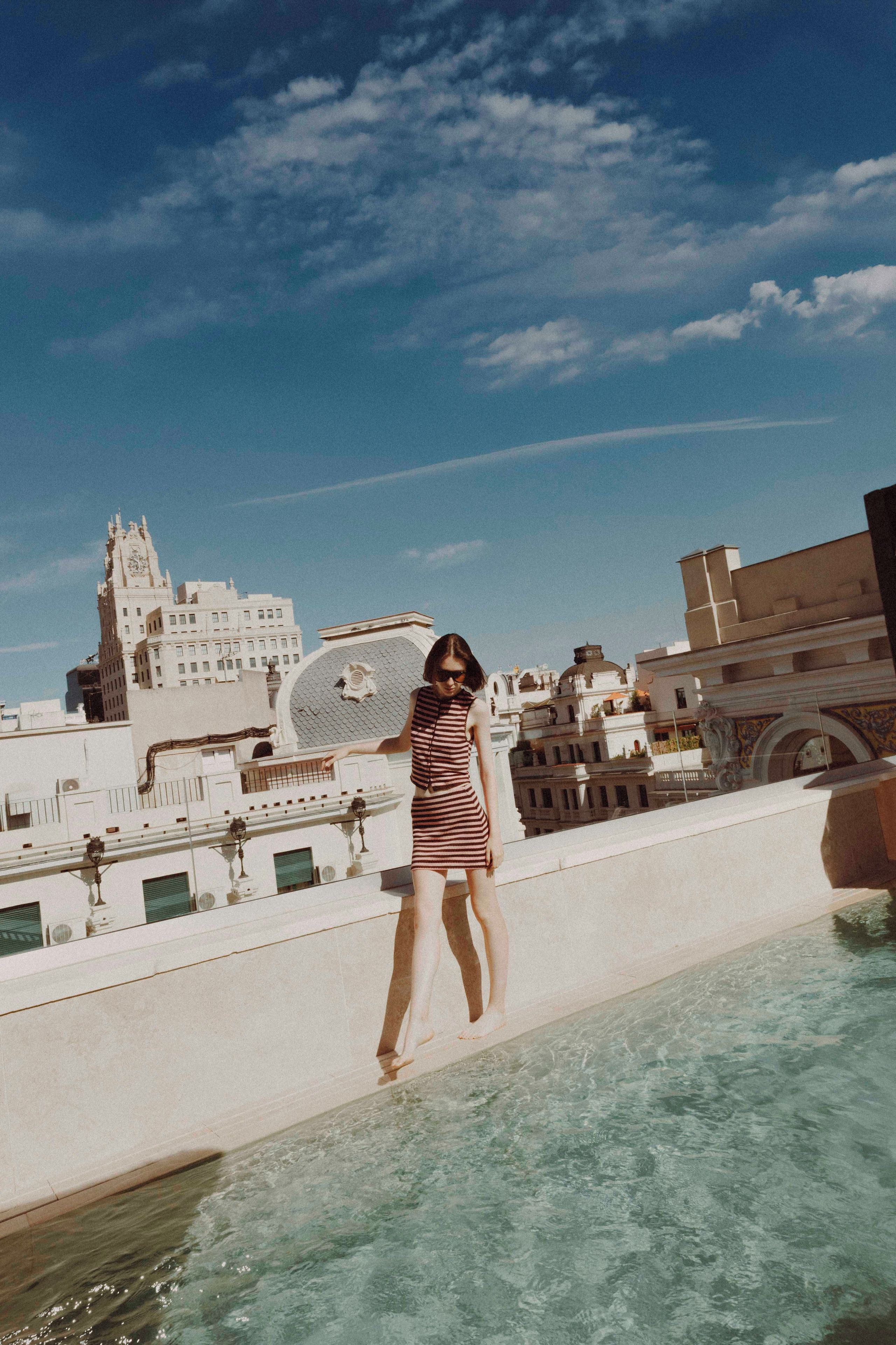 Una persona de pie junto a una piscina, con un paisaje urbano al fondo.