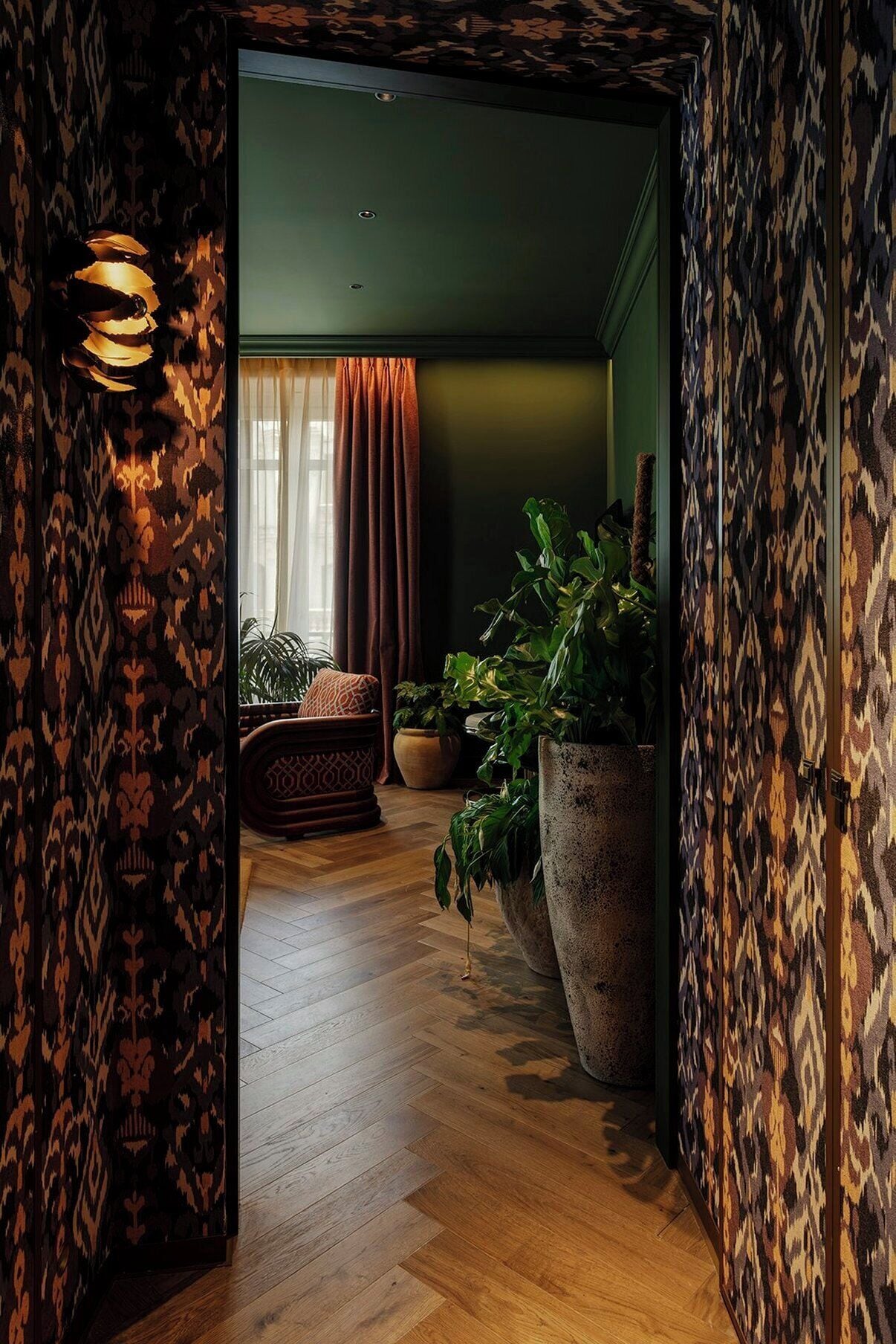 A hallway with patterned wallpaper, a wooden floor, a large potted plant, a patterned sofa, and a lamp.
