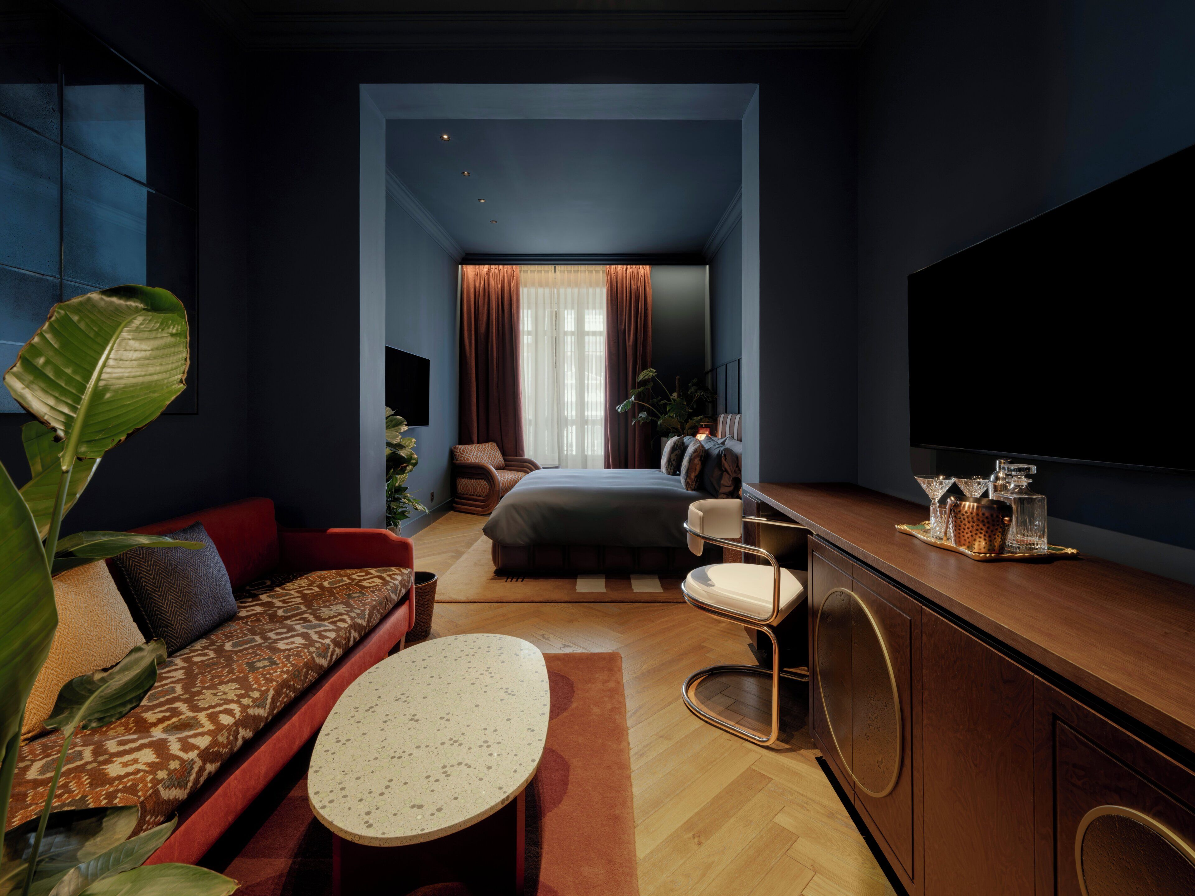 A modern hotel room with a large bed, a seating area with a sofa and armchair, a small round table, and a television on the wall.