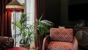 Un salón con un sofá rojo, un sillón estampado y una pequeña mesa redonda. Hay plantas en macetas y un ventanal con cortinas.