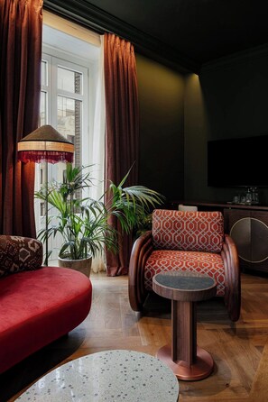 A living room with a red sofa, patterned armchair, and a small round table. There are potted plants and a large window with curtains. - Nômade Temple Madrid (Madrid)