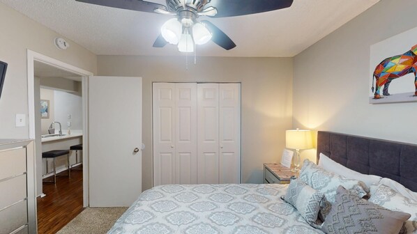 A bedroom with a bed, a nightstand, a lamp, a ceiling fan, a wardrobe, and a bathroom with a sink and mirror.