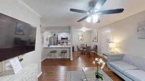 A modern living room with a flat-screen TV, a ceiling fan, a dining area with a table and chairs, and a kitchen in the background.