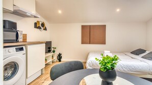 A compact hotel room with a kitchenette, a bed, a small table with a plant, and a wall-mounted TV. - Le Factory - Studio contemporain - Proche métro B (Oullins-Pierre-Bénite)