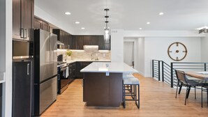 A modern kitchen with dark cabinetry, stainless steel appliances, a central island with bar stools, and a dining area with a wooden table and chairs. - New Ski/Summer Home (Cottonwood Heights)