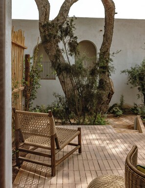 Une chaise en osier et une petite table ronde dans une cour, avec un arbre et un mur blanc.