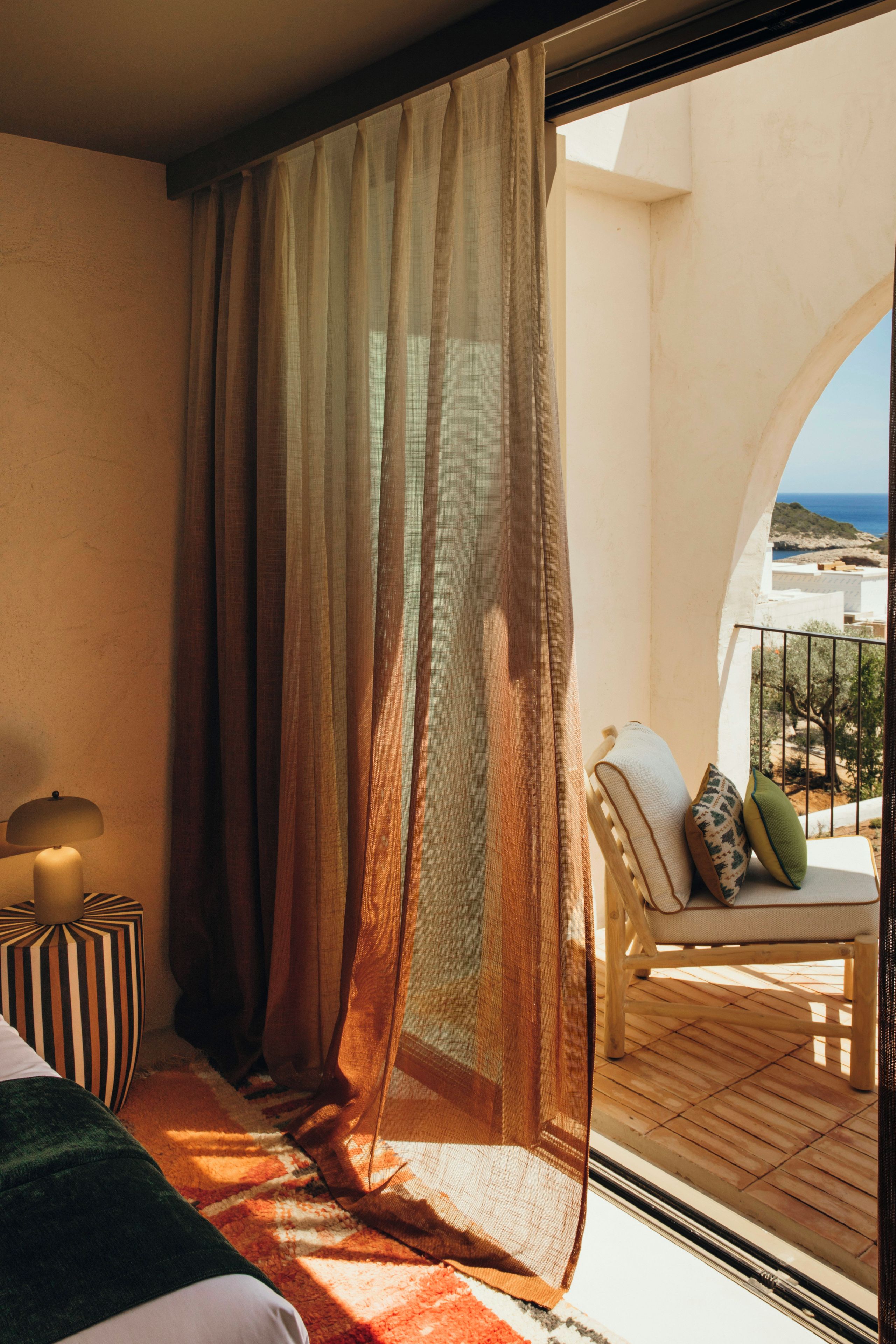 Un balcón con vista al mar, una silla con cojines y una cama con almohada a rayas.