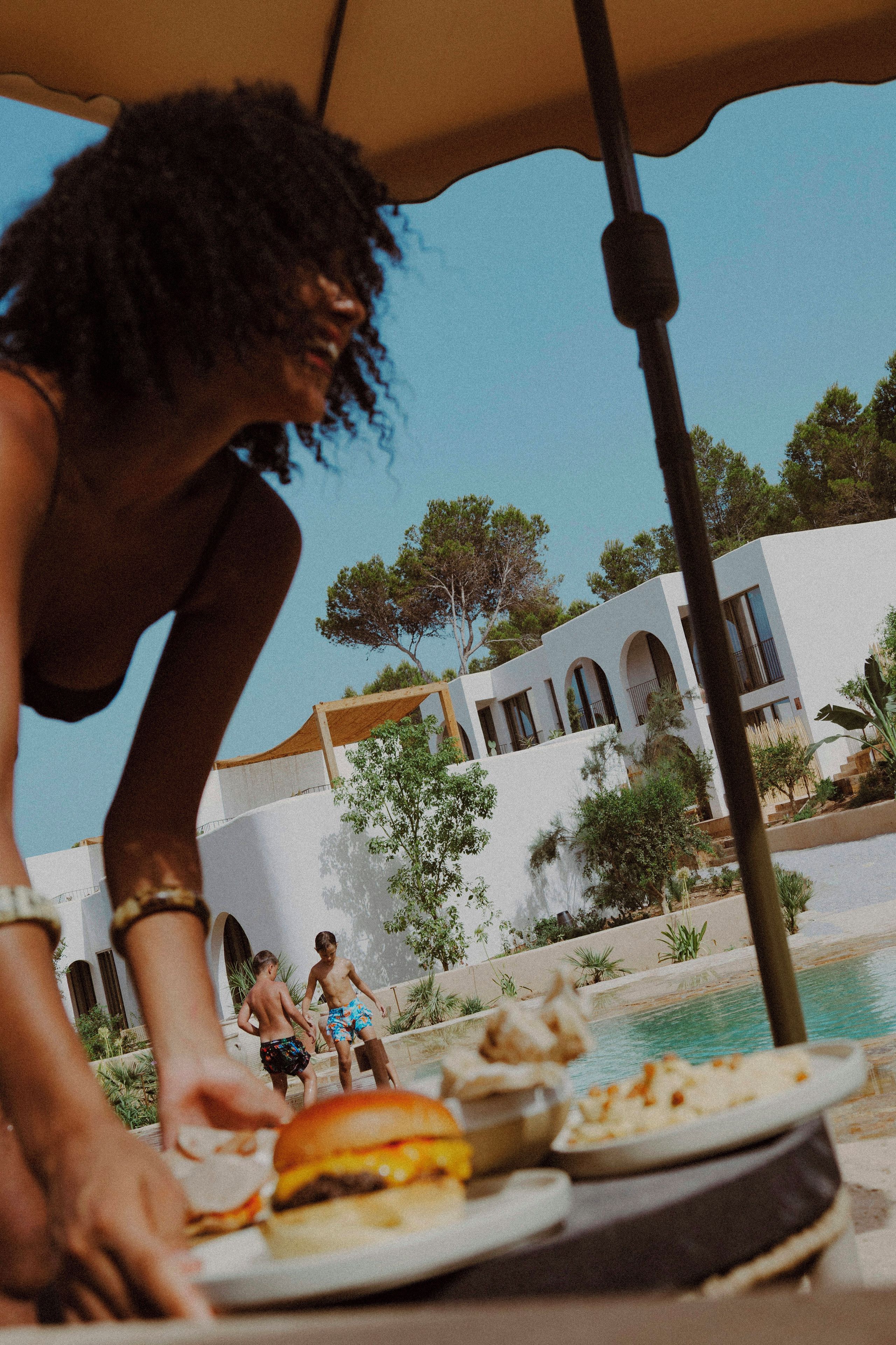 Un área junto a la piscina con mesa de comedor, piscina y un edificio con arcos al fondo.