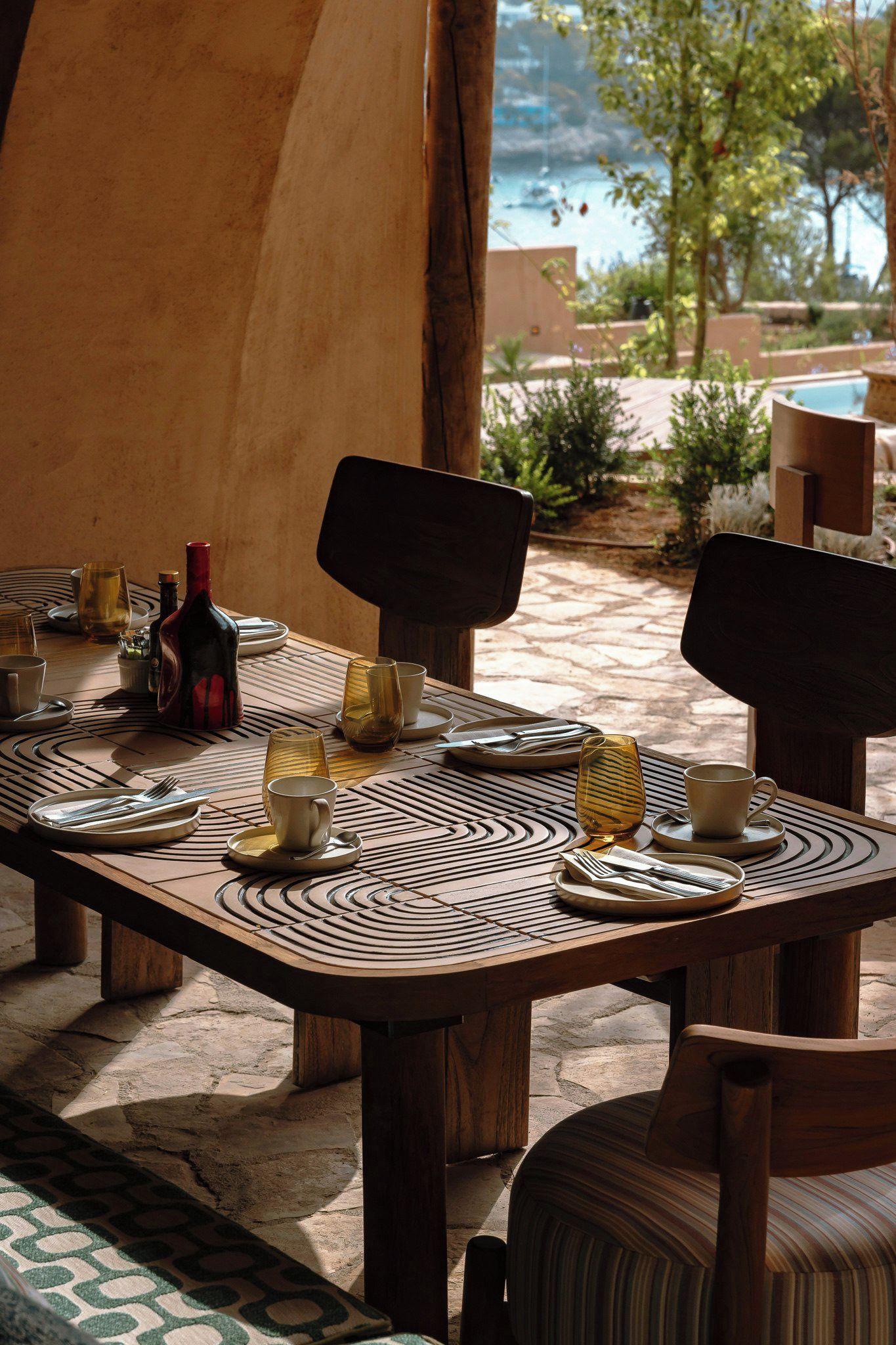An outdoor dining area with a table set for two, surrounded by chairs, and a view of a pool and greenery.