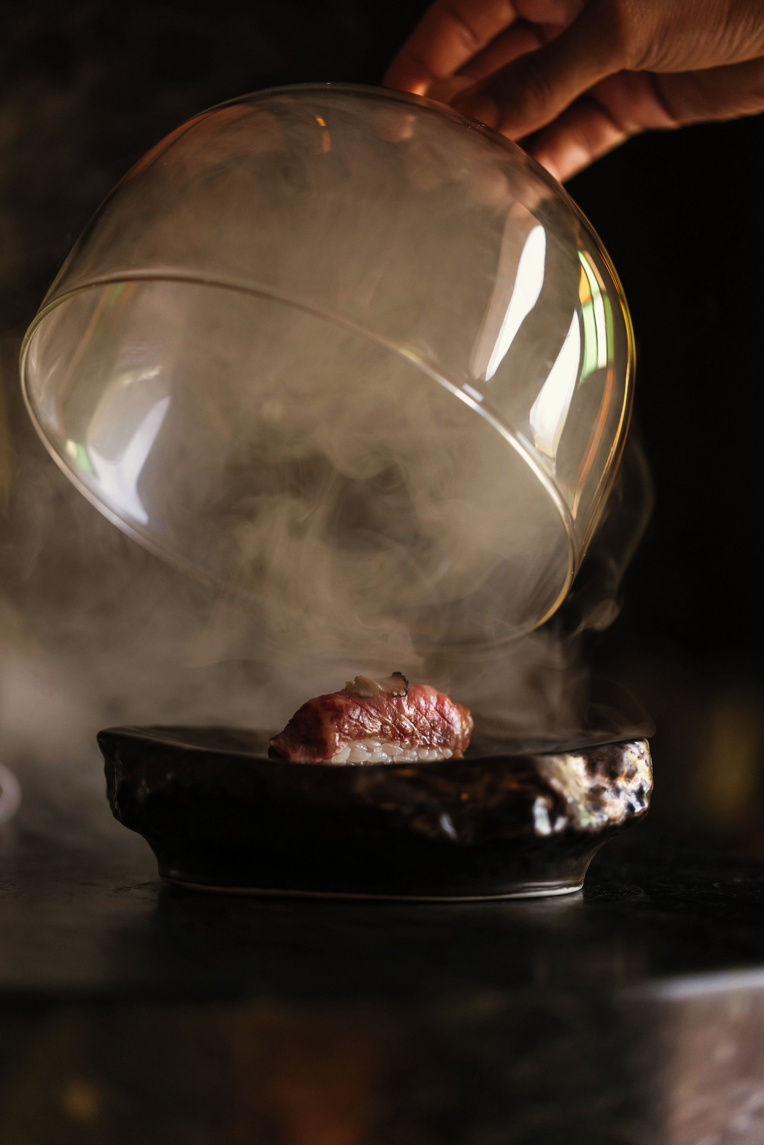 A dome-shaped cover over a dish with food, steam rising from it, and a hand adjusting the cover.