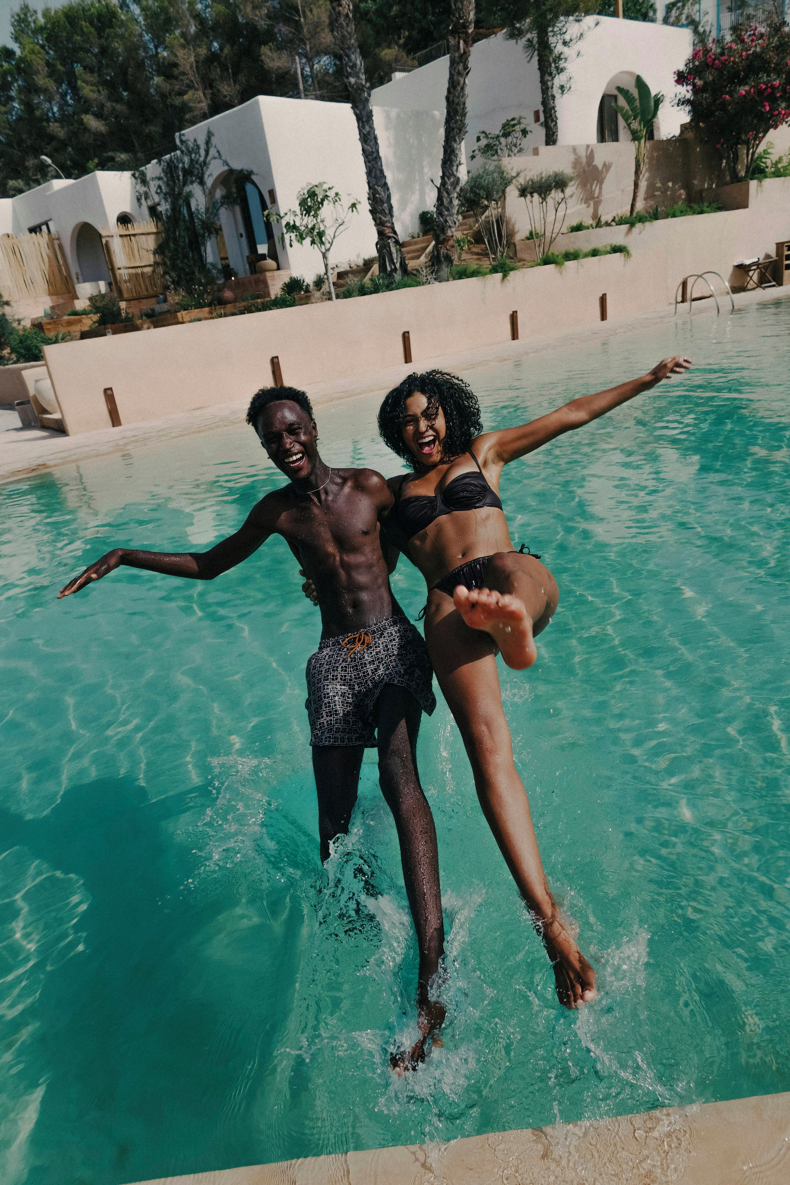 Due persone in piscina con acqua cristallina, circondate da un edificio bianco con archi e vegetazione.