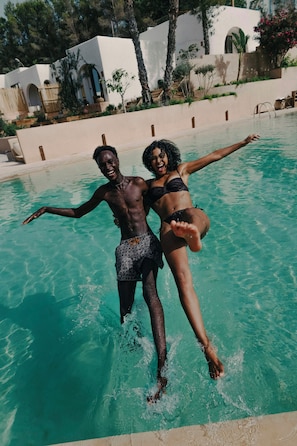 Dos personas en una piscina con agua cristalina, rodeadas por un edificio blanco con arcos y follaje.