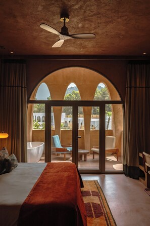 A bedroom with a bed, a ceiling fan, and a view through an arched window to an outdoor seating area.