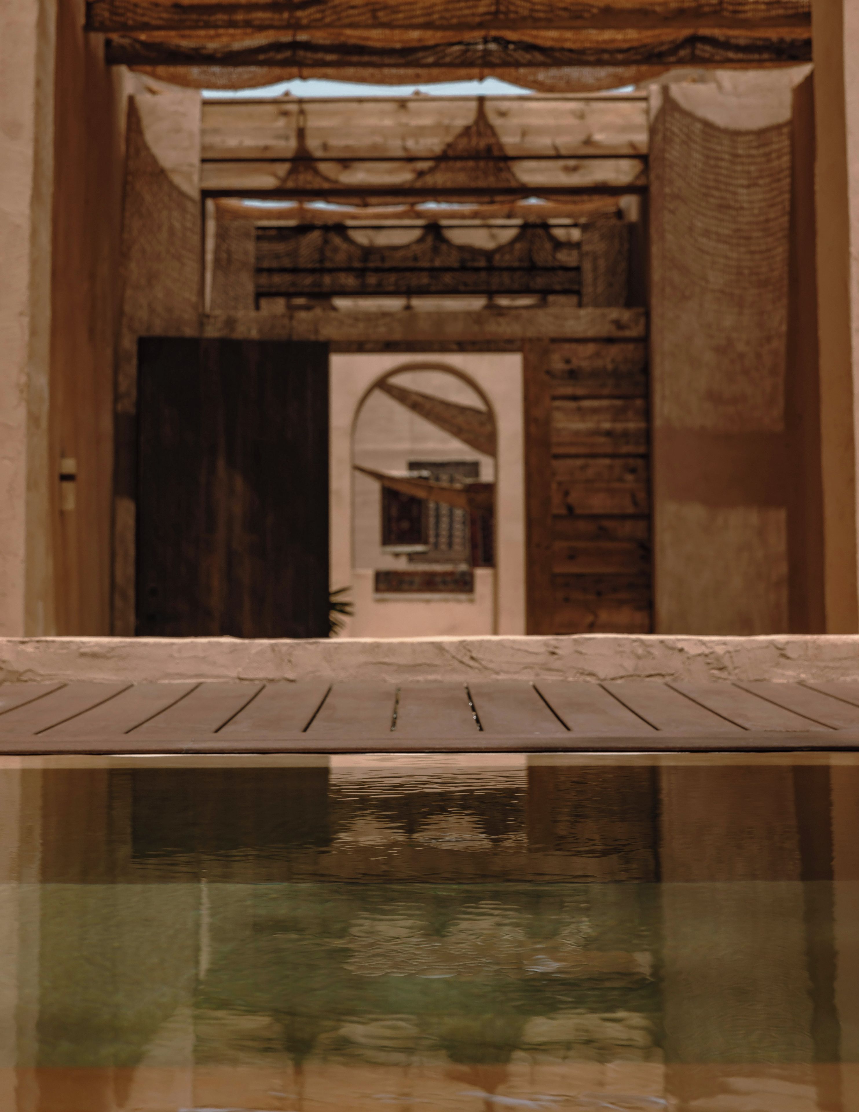 Ein Pool mit einer Holzterrasse im Vordergrund und Blick auf ein Gebäude mit Rundbogenfenstern und Holzbalken im Hintergrund.