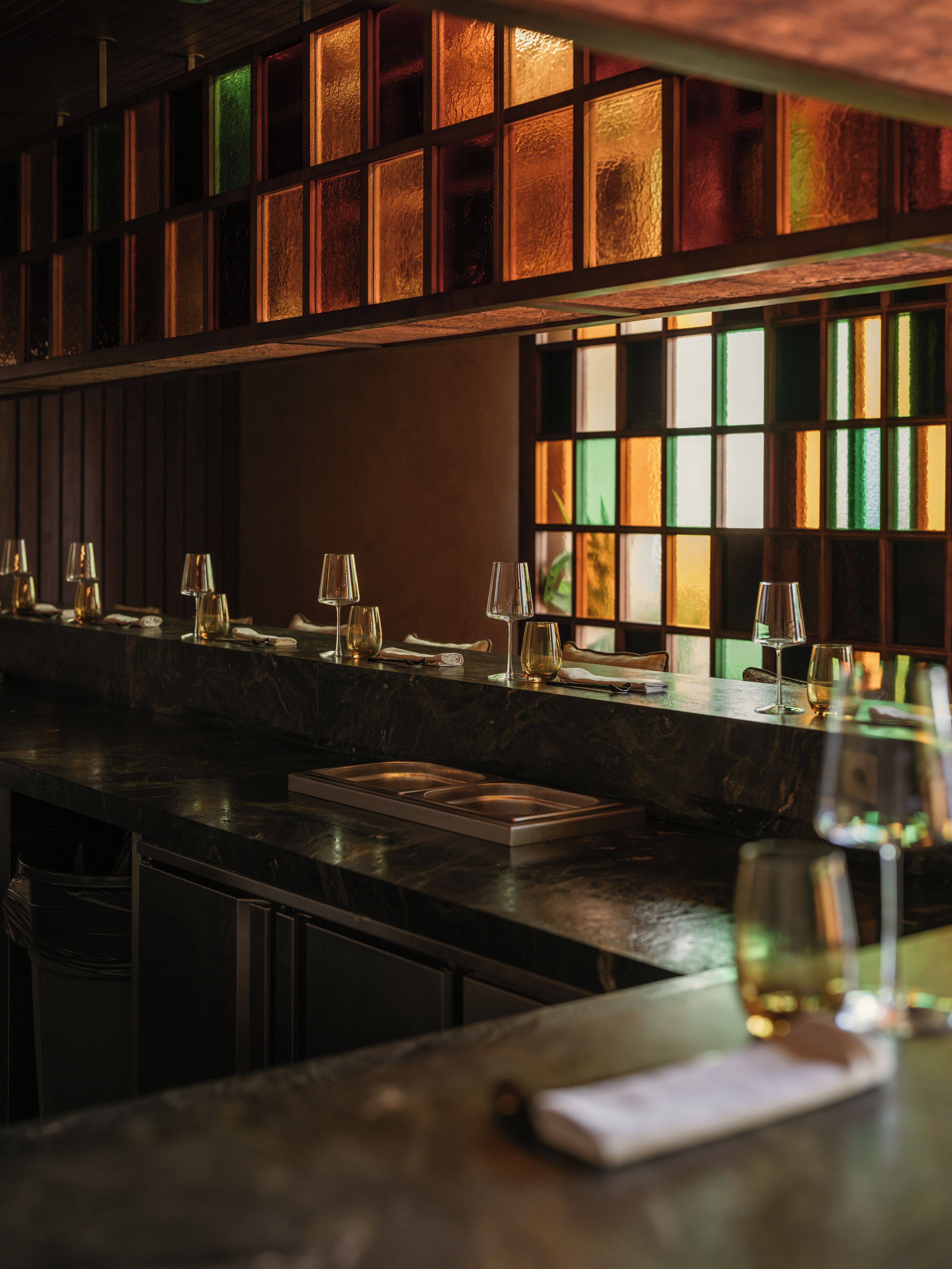 Un espace bar doté d’un comptoir sombre, de nombreux verres et d’une fenêtre ornée de vitraux colorés.