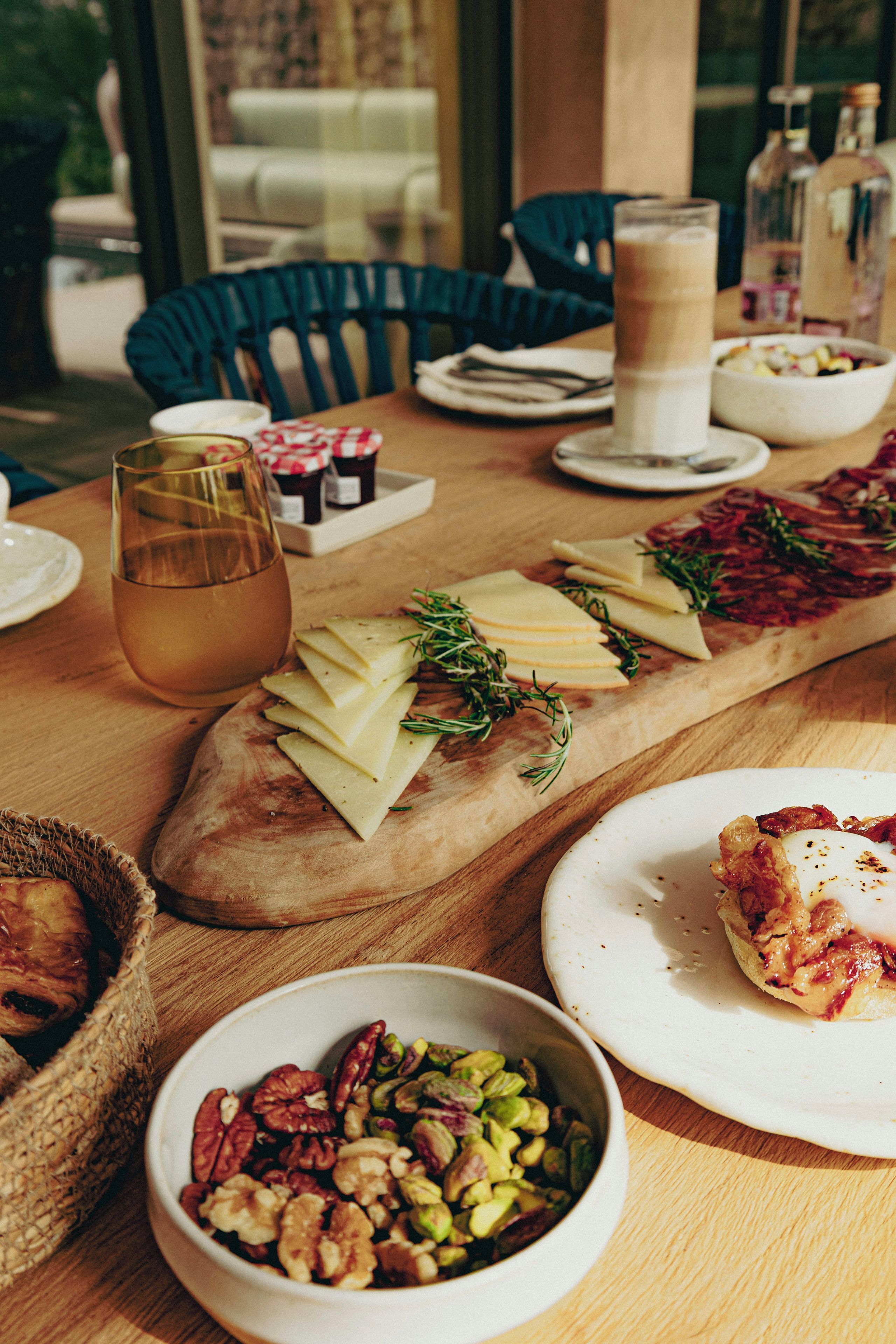 Una mesa dispuesta con queso, frutos secos y una bebida, con sillas y botellas al fondo.