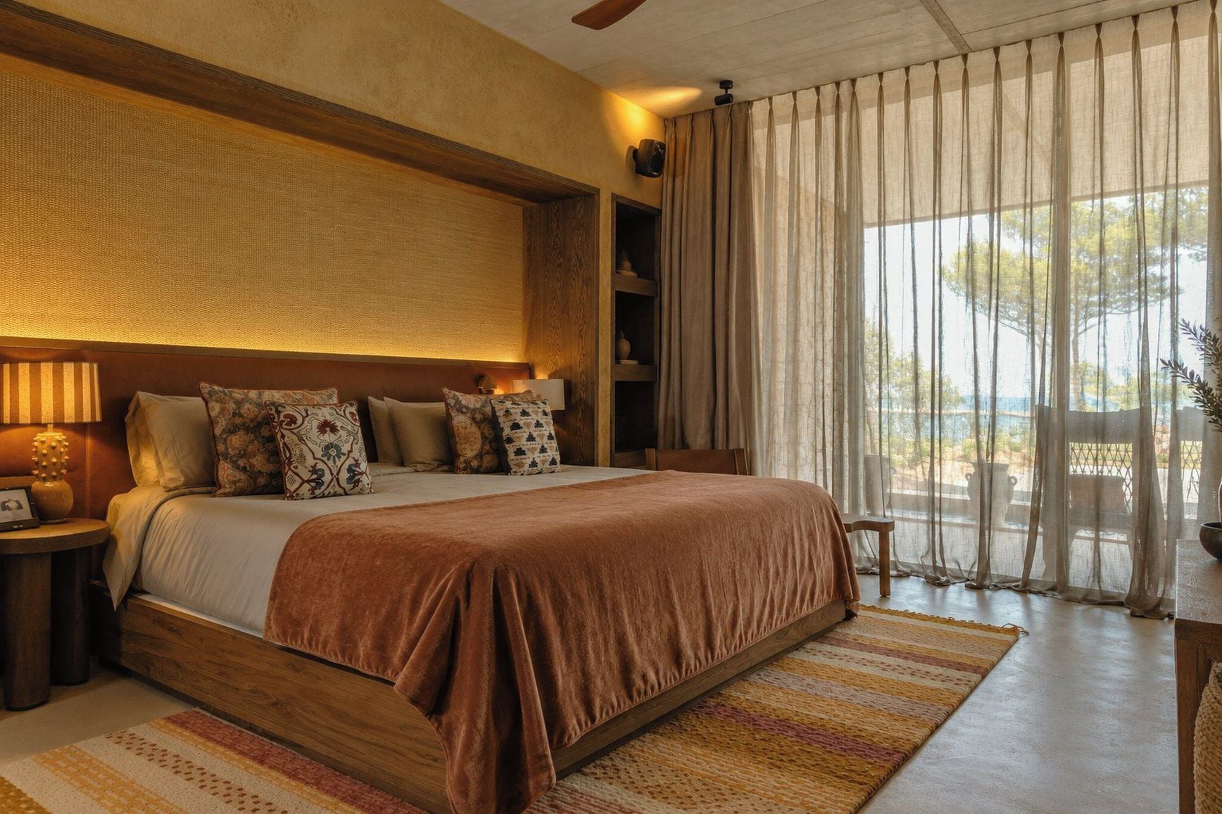 Un dormitorio amplio con una cama grande, cabecera de madera, mesita de noche con lámpara y vistas al exterior a través de cortinas traslúcidas.
