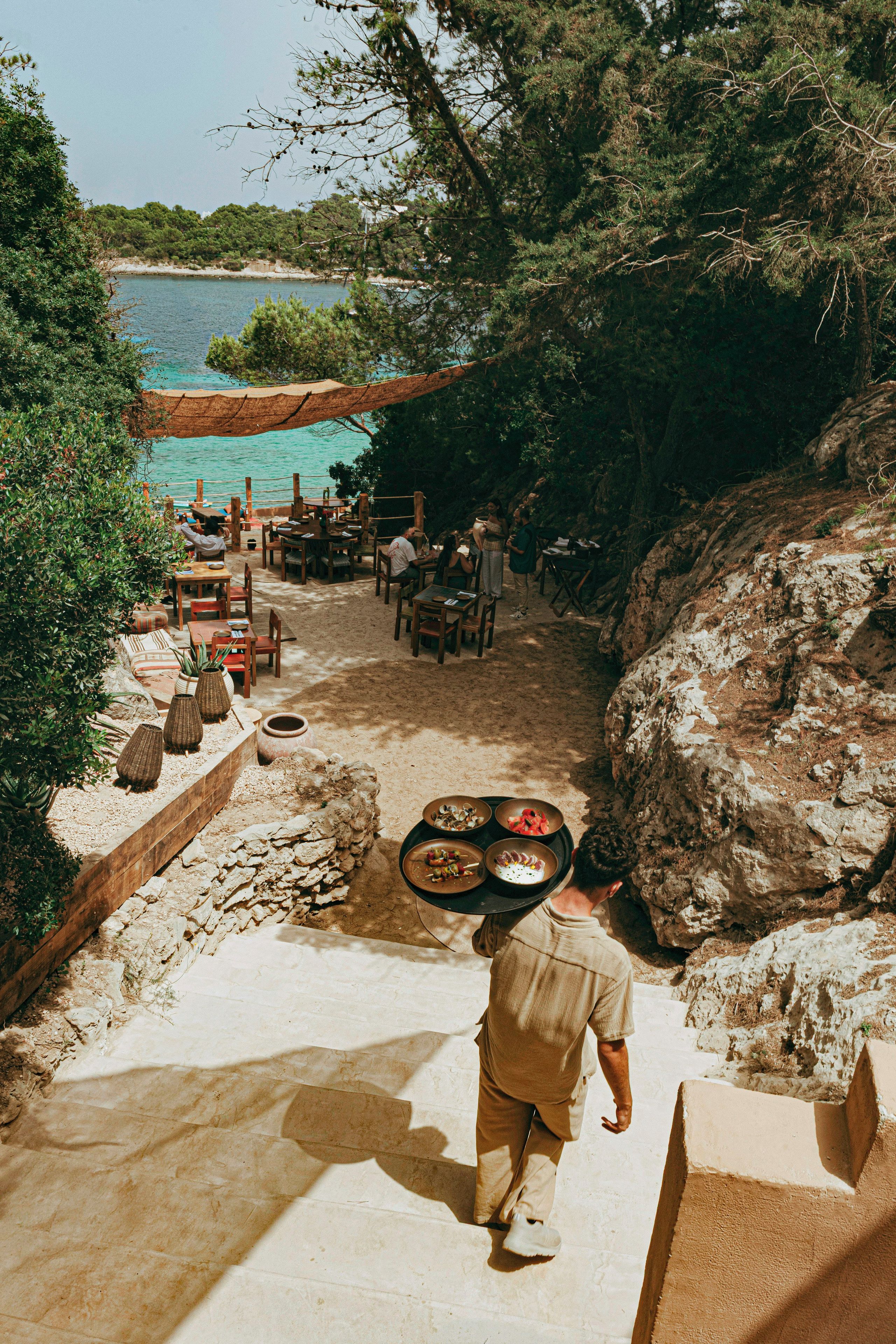 A person carrying a tray of food up a stone staircase to a dining area with tables and chairs, overlooking a body of water.