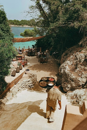 Una persona lleva una bandeja con comida por una escalera de piedra hacia un área de comedor con mesas y sillas, con vista a una masa de agua.