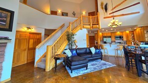 A spacious living area with a black leather sofa, wooden staircase, and a kitchen area.