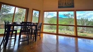 A dining area with a table and chairs, large windows with a view of a forested landscape, and a painting on the wall.