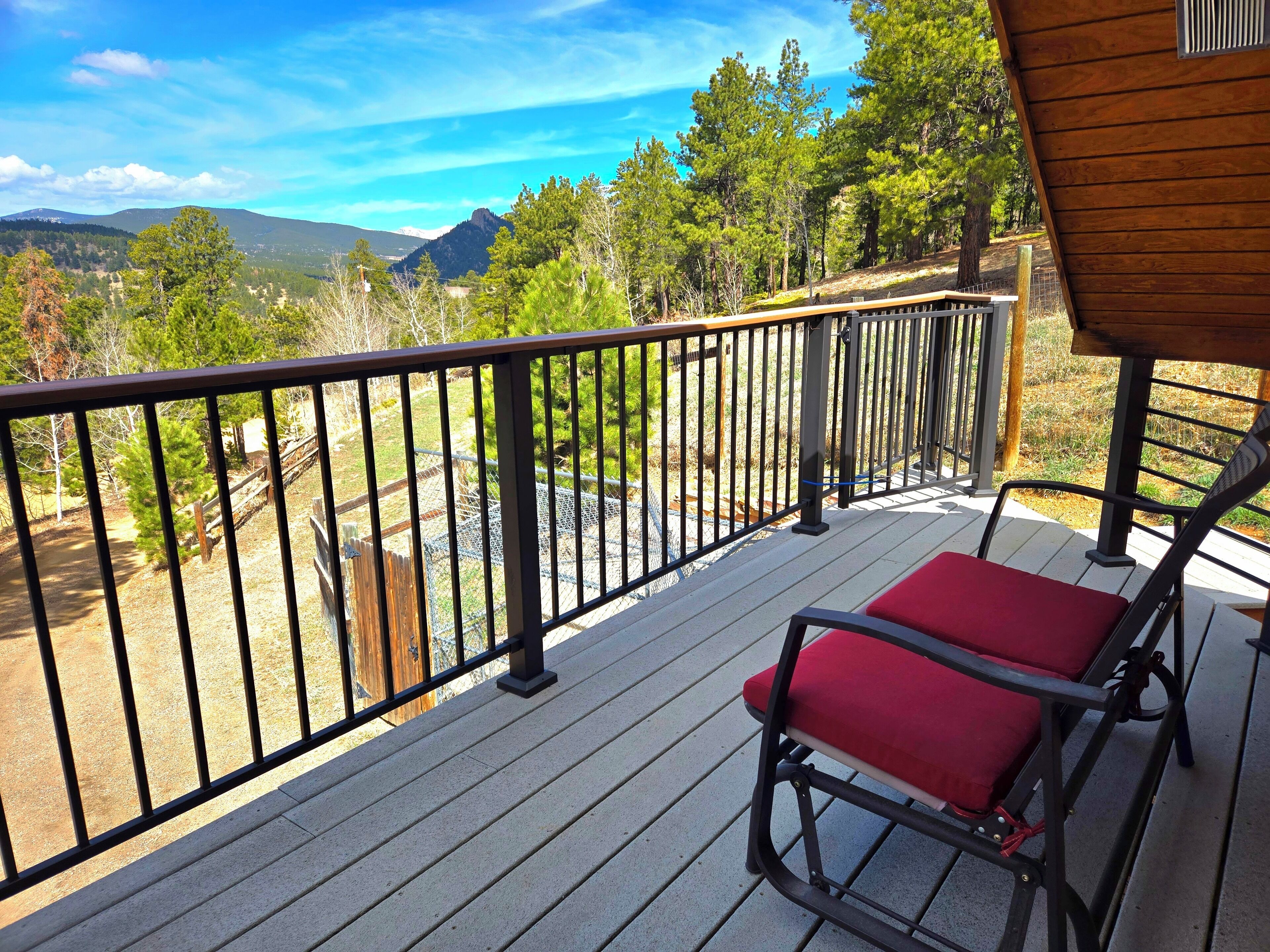 Una terraza de madera con barandilla metálica, dos sillas rojas con cojines y vistas a un paisaje montañoso.