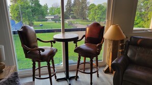 Una habitación con dos sillas de madera, una mesita, una lámpara y vistas a un parque a través de un ventanal grande.