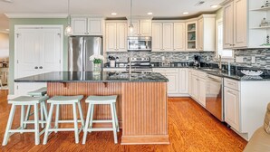 A modern kitchen with a central island, bar stools, stainless steel appliances, and white cabinetry. - Walk to Beach! Sunny 5BR Oak Island Oasis (Oak Island)