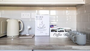 A kitchen counter with a coffee station, including a kettle, mugs, and a sign offering complimentary tea, coffee, and biscuits. - London, Heathrow & Twickenham Stadium Stay (Isleworth)