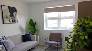 A living room with a sofa, a chair, a large plant, and a window with blinds. - London, Heathrow & Twickenham Stadium Stay (Isleworth)