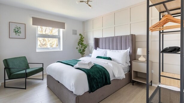 A modern bedroom with a bed, a green chair, a bedside table with a lamp, and a window with a view of trees. - London, Heathrow & Twickenham Stadium Stay (Isleworth)