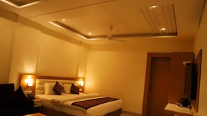 A hotel room with a bed, bedside lamps, a desk, a chair, a TV, a ceiling fan, and a door. - Ananda Castle (Vrindavan)