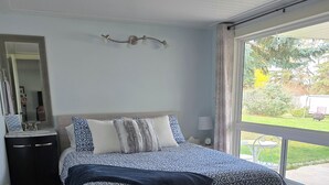 A bedroom with a bed, a nightstand, a mirror, and a window with a view of the outdoors.