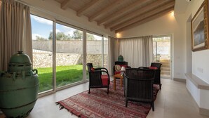 Una habitación amplia con grandes puertas correderas de vidrio que dan a un jardín, muebles de mimbre, una maceta verde grande y un cuadro en la pared.
