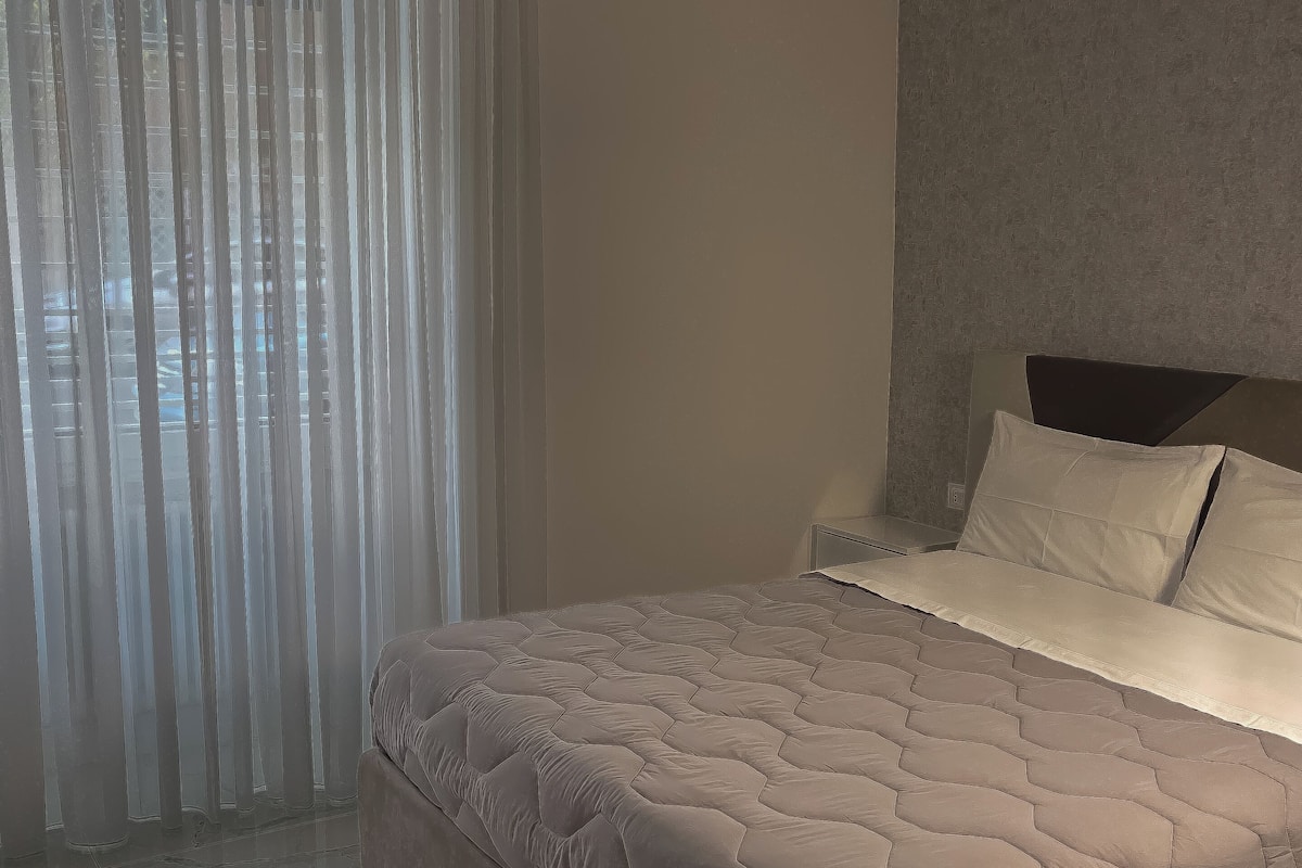 A bedroom with a bed, a headboard, and a window with sheer curtains.