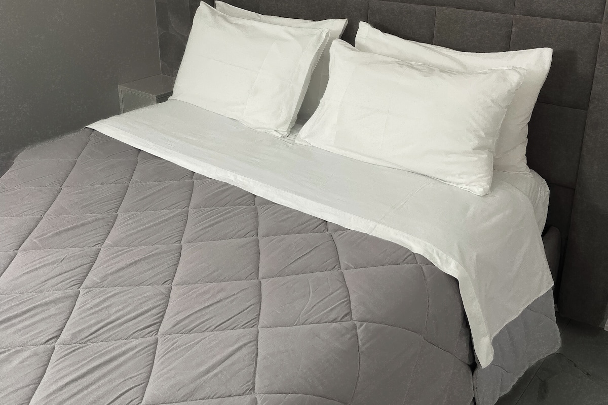 A bed with a quilted grey mattress and white pillows, set against a textured grey wall.
