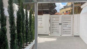 An outdoor covered patio area with a tiled floor, white metal gates, and a row of evergreen trees. - Alissa Suite (Rome)