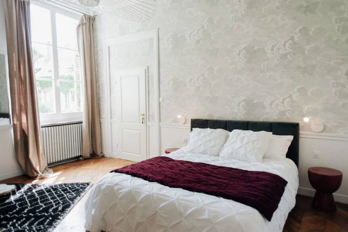 Ein Schlafzimmer mit Bett, Nachttisch, Fenster mit Vorhängen und Heizkörper.