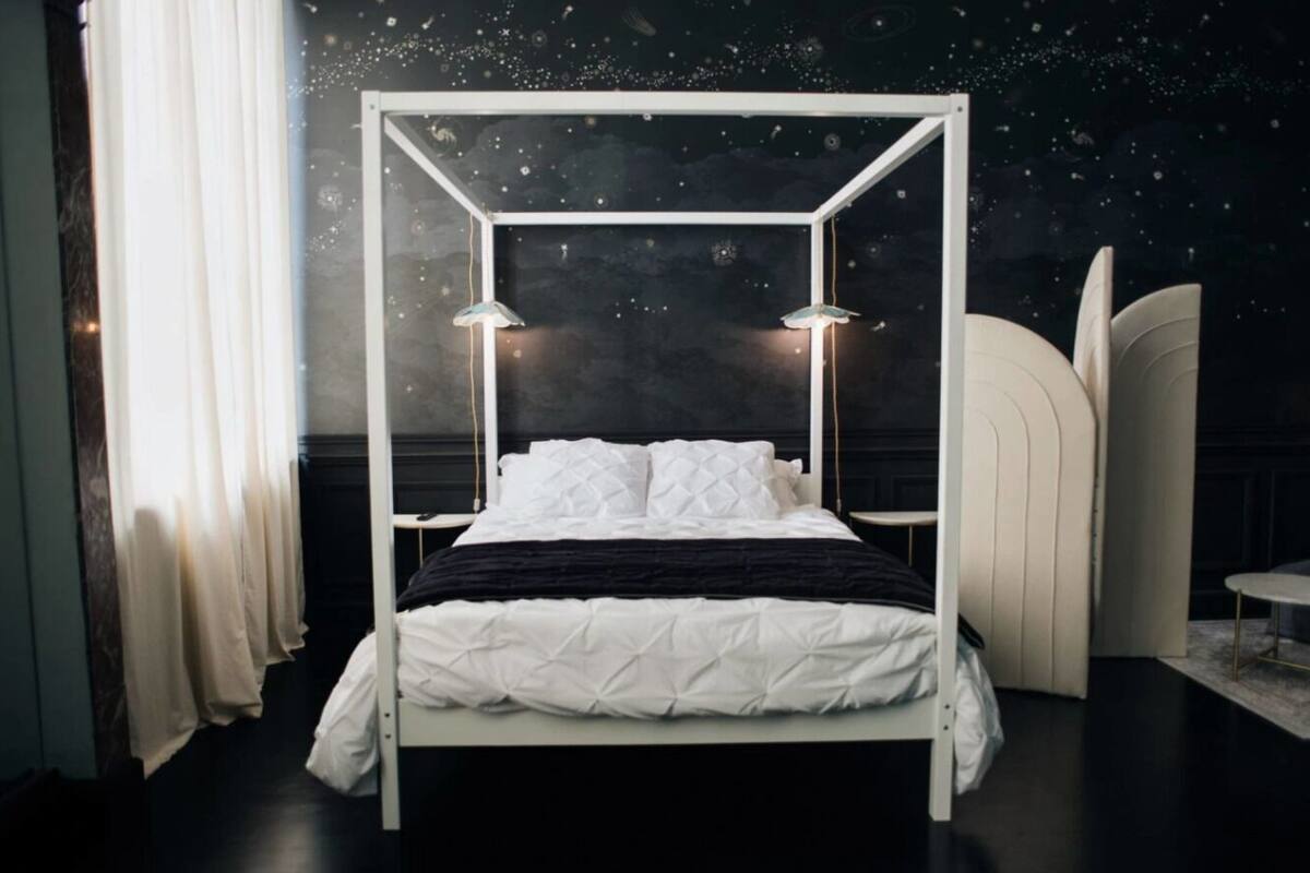 Ein Himmelbett mit weißem Gestell und schwarzer Bettwäsche, zwei Nachttischlampen und ein Wandgemälde mit Sternenmotiven.