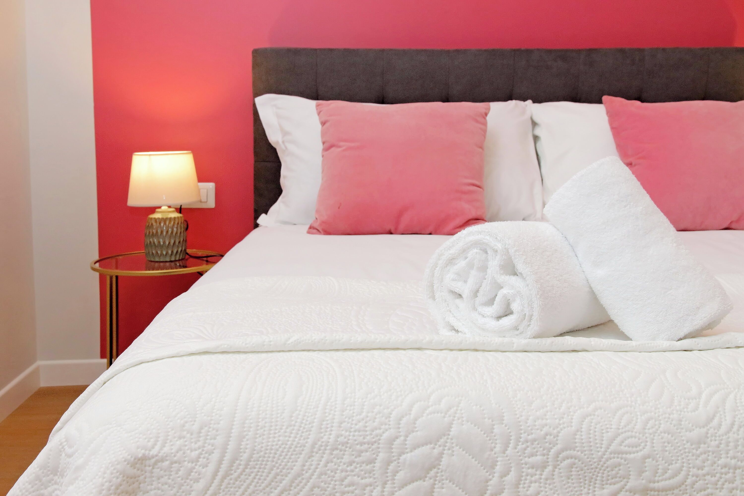Un lit bien fait, avec une literie blanche et des oreillers roses, une lampe de chevet et un mur rouge.
