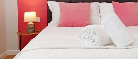 Un lit bien fait, avec une literie blanche et des oreillers roses, une lampe de chevet et un mur rouge.