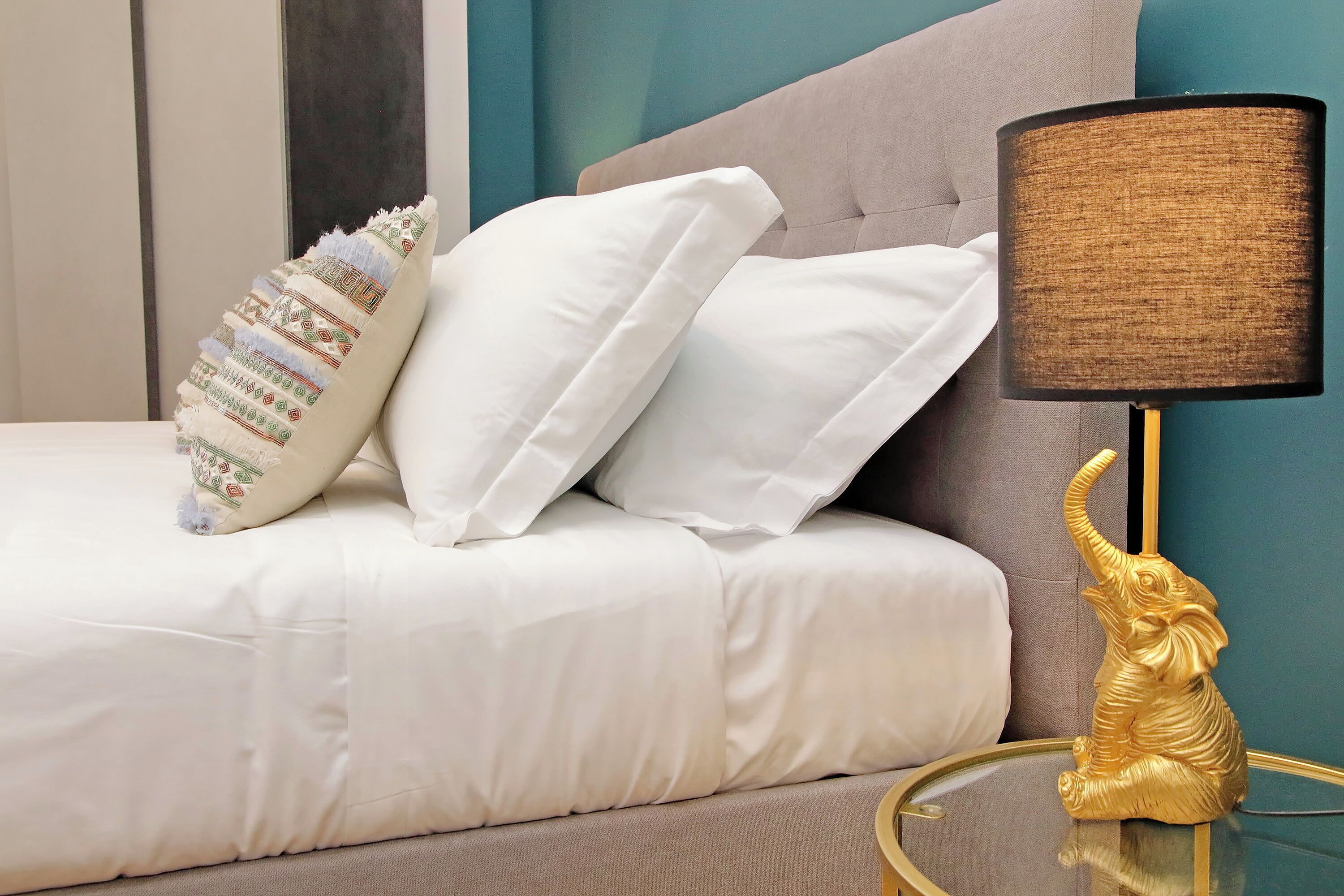 A bed with white and patterned pillows, a beige headboard, and a gold elephant lamp on a glass table.